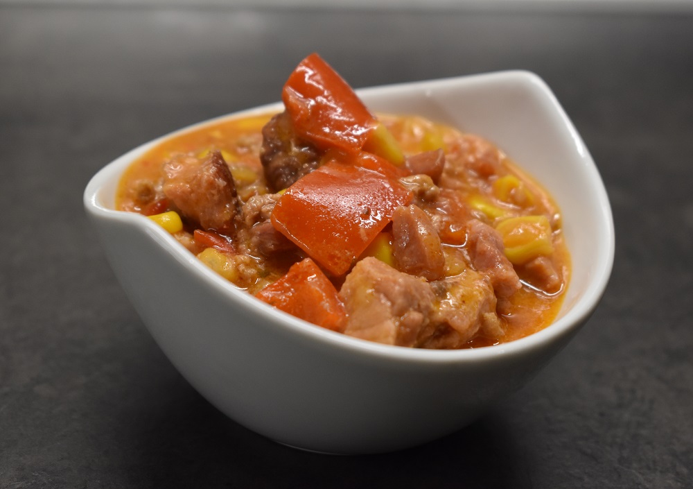 Eine weiße Schale, gefüllt mit einem stückigen Eintopf, der Fleischstücke, rote Paprika, Mais und Tomaten in einer dicken, rötlichen Brühe enthält und auf einer dunklen Oberfläche steht.