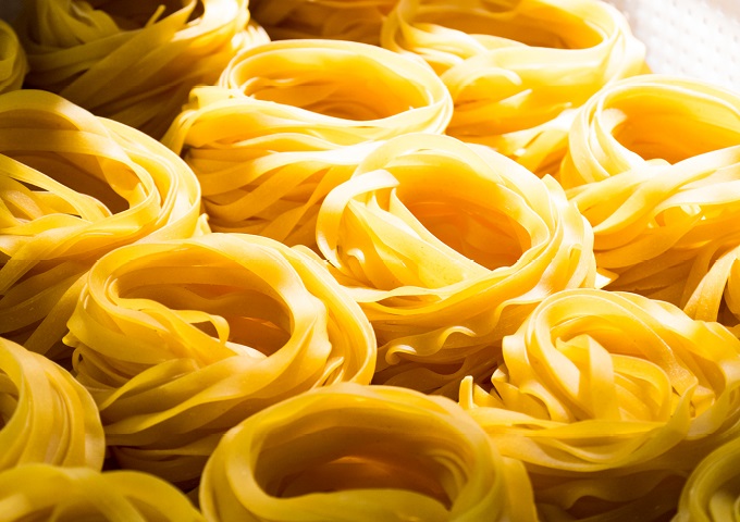 Nahaufnahme von mehreren Nestern ungekochter gelber Tagliatelle-Nudeln, die dicht aneinander gestapelt und von hellem Licht beleuchtet sind.