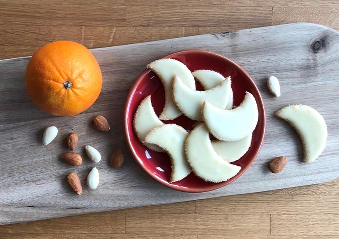 Ein Holzbrett mit einer Orange, verstreuten Mandeln und einem roten Teller mit halbmondförmigen weißen Keksen aus Zuckerguss, neben dem einige Kekse und Mandeln liegen.