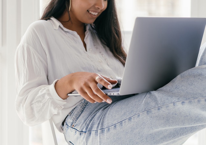 Eine Frau in weißer Bluse und hellblauen Jeans sitzt mit übereinandergeschlagenen Beinen da und lächelt, während sie einen Laptop auf ihrem Schoß benutzt. Der helle Innenraum spiegelt die Idee von "Vorsorgen statt verzichten" wider - im Voraus planen statt verzichten.
