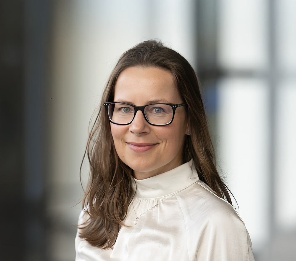 Eine Frau mit langen braunen Haaren und Brille, die eine weiße Bluse trägt, lächelt in die Kamera. Der Hintergrund ist mit natürlichem Licht und Fensterelementen sanft verschwommen.