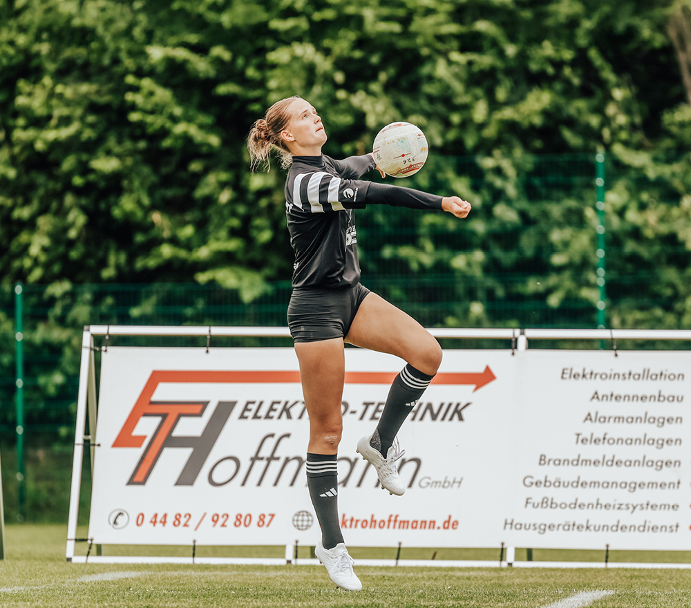 Eine Sportlerin in einem schwarzen Trikot springt, um während eines Sportspiels im Freien einen Ball mit ihren Unterarmen zu treffen. Im Hintergrund sind ein Werbebanner eines Unternehmens und grüne Bäume zu sehen.