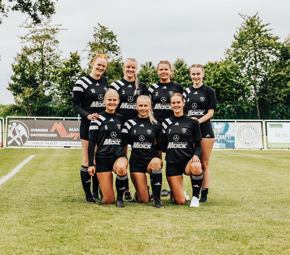 Sieben Frauen in passenden schwarzen Fußballtrikots posieren gemeinsam auf einer Wiese, vier davon stehend und drei kniend vor ihnen. Im Hintergrund sind Bäume und Werbeschilder zu sehen.