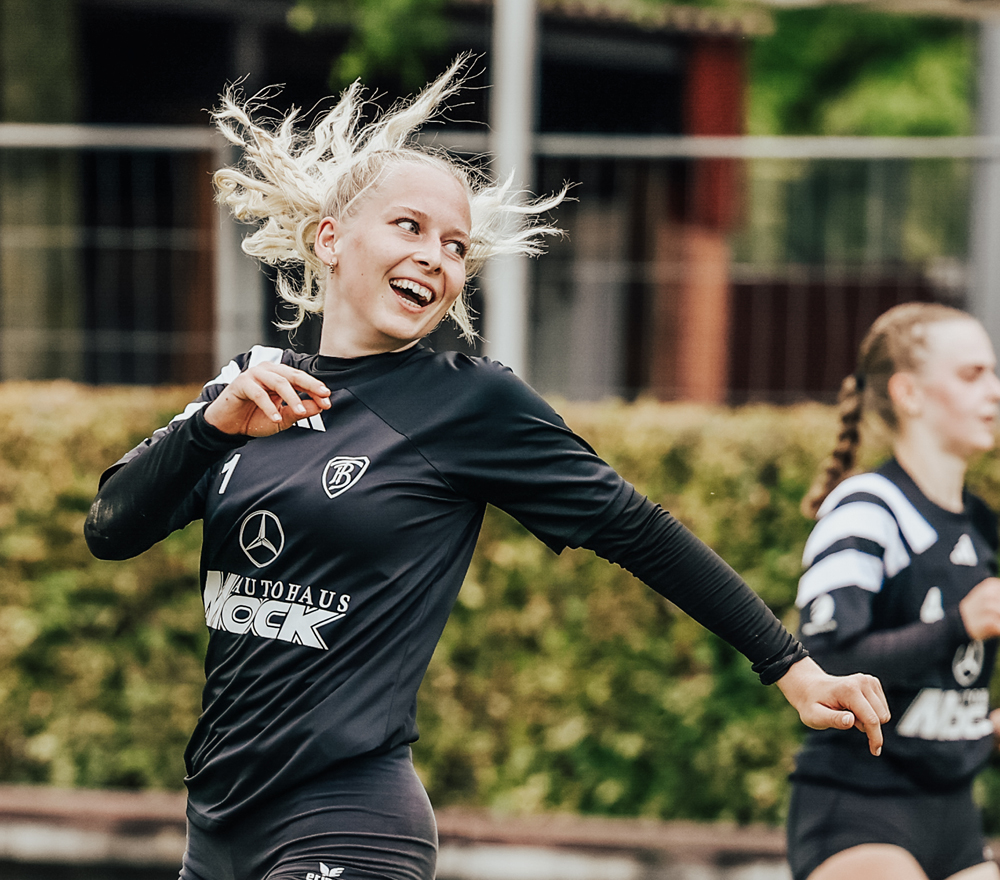 Eine Fußballspielerin mit blonden Haaren in einem Pferdeschwanz lächelt, während sie während eines Spiels in einem schwarzen Trikot mit Sponsorenlogos läuft. Ein anderer Spieler in ähnlicher Kleidung ist im Hintergrund unscharf zu erkennen.
