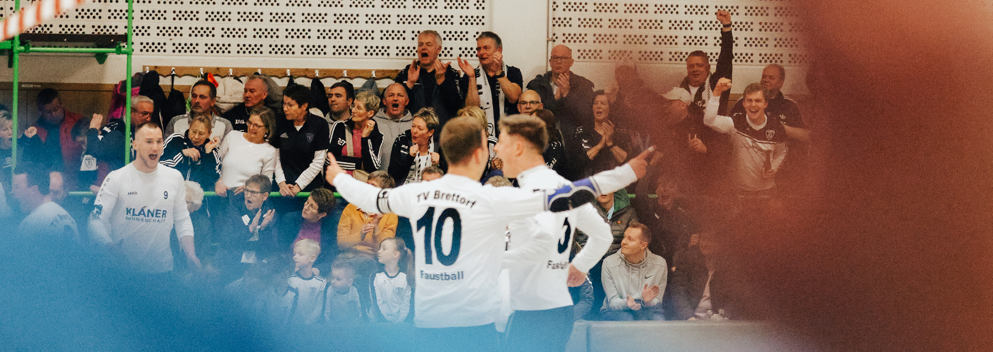 Hallenfußballspieler in weißen Trikots jubeln mit ausgestreckten Armen auf dem Spielfeld, während eine große Menge von Fans im Hintergrund von der Tribüne aus begeistert anfeuert.
