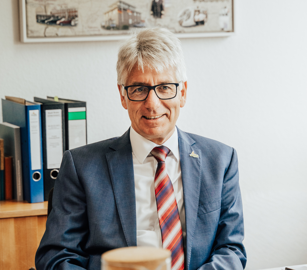 Ein Mann mittleren Alters mit grauem Haar und Brille, der einen blauen Anzug, ein weißes Hemd und eine rot gestreifte Krawatte trägt, sitzt an einem Schreibtisch in einem Büro mit Ordnern und gerahmten Bildern im Hintergrund.