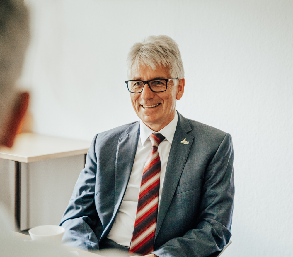 Ein lächelnder Mann im Anzug und mit rot gestreifter Krawatte sitzt an einem Schreibtisch, einer anderen Person gegenüber, die mit dem Rücken zur Kamera steht, in einer hellen, professionellen Büroumgebung.