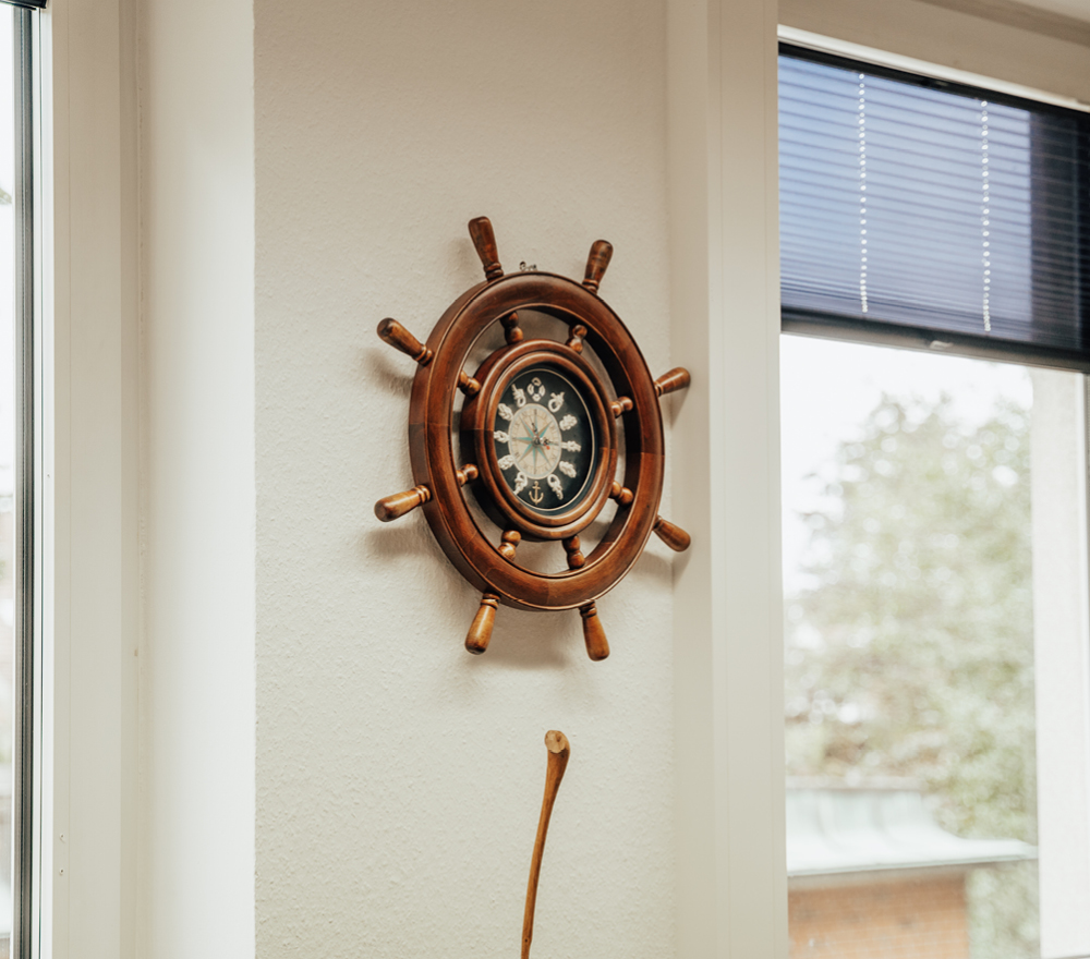 Eine Wanduhr in Form eines Schiffsrades hängt an einer weißen Wand zwischen zwei Fenstern mit Jalousien, durch die natürliches Licht einfällt. Durch das Fenster sind draußen Bäume und ein Gebäude zu sehen.