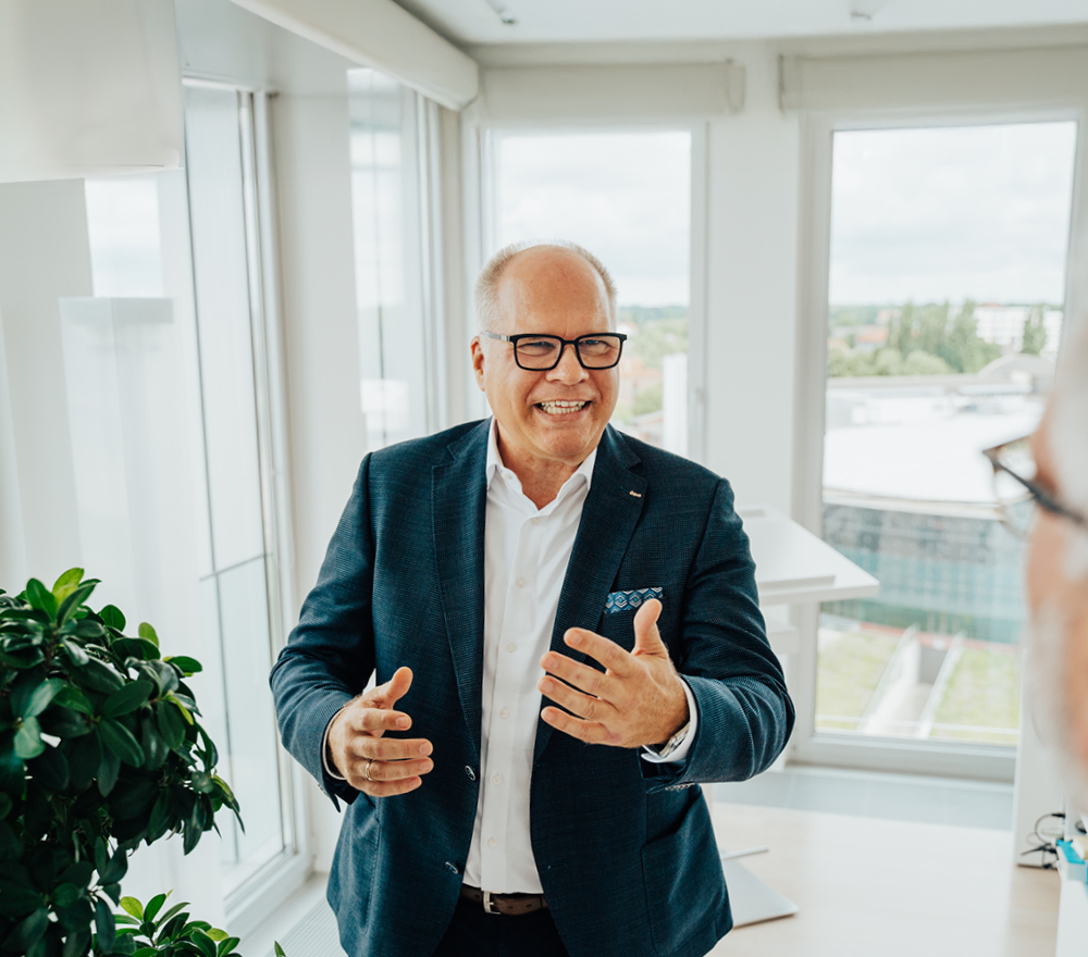 Ein lächelnder älterer Mann im Anzug und mit Brille gestikuliert, während er in einem hellen, modernen Büro mit großen Fenstern steht und eine Topfpflanze neben sich sieht.