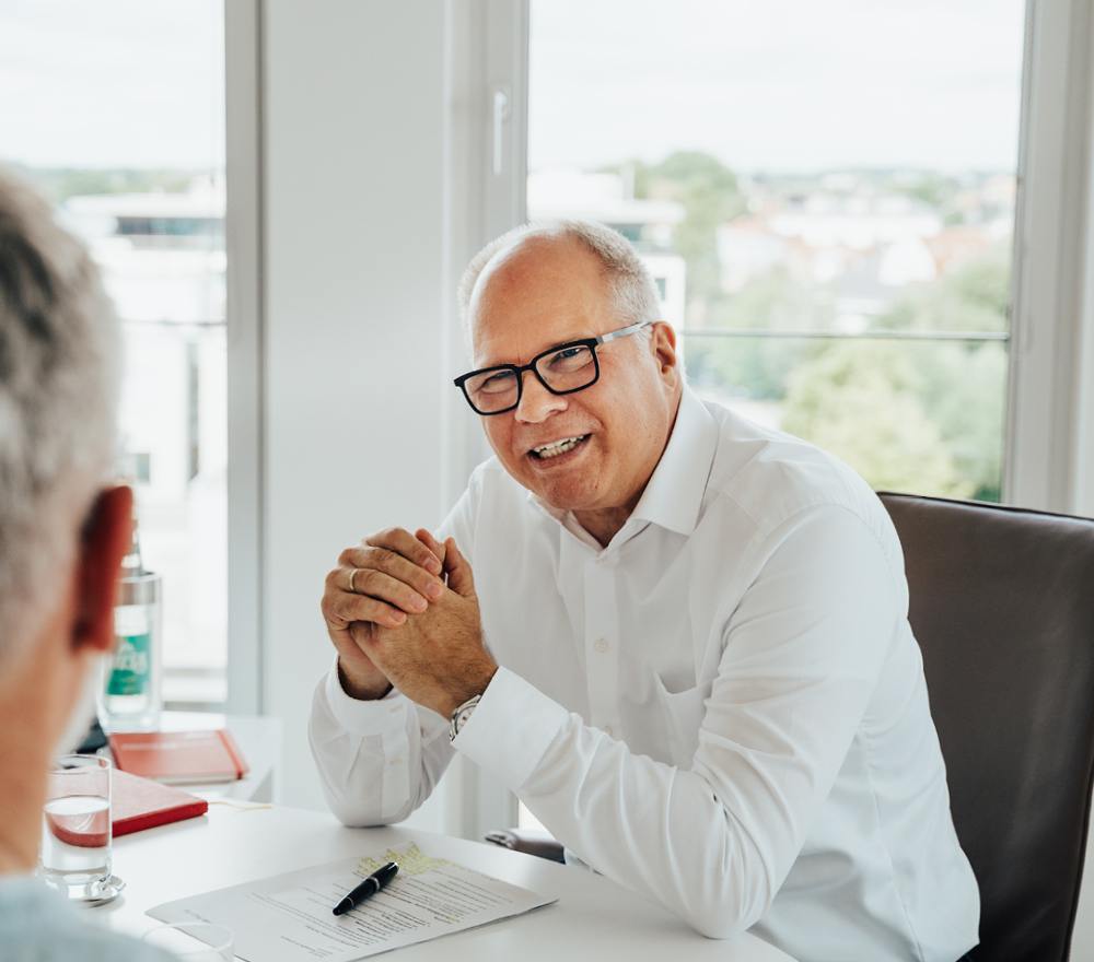 Ein älterer Mann mit weißem Hemd und Brille lächelt, während er an einem Schreibtisch mit Dokumenten, einem Stift und einem Glas Wasser sitzt und sich mit einer anderen Person in einem hellen Büro unterhält.