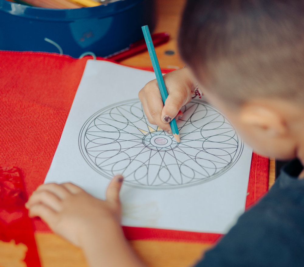 Ein Kind malt mit einem blauen Farbstift ein geometrisches Mandala auf ein Blatt Papier, während es an einem Tisch mit einer roten Matte und einer Schachtel mit Farbstiften sitzt.