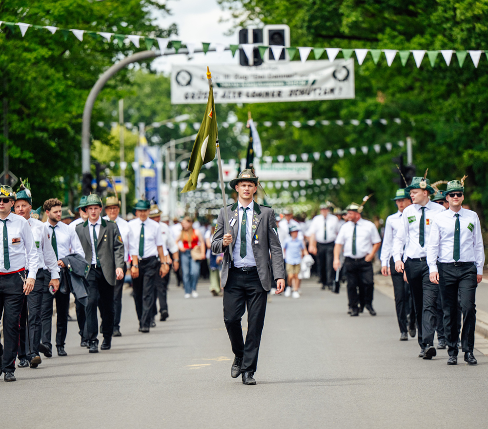 Eine Gruppe von Männern in weißen Hemden, schwarzen Hosen und Hüten mit grünen Verzierungen marschiert eine von Bäumen gesäumte Straße entlang, angeführt von einem Mann mit einer Flagge. Wimpel und Fahnen hängen über dem Kopf.
