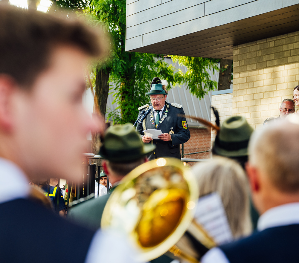 Ein Mann in grüner Uniform und Hut spricht im Freien an einem Mikrofon und hält Papiere in der Hand. Im Vordergrund sind Musiker, einige mit Blasinstrumenten, zu sehen, deren Gesichter verschwommen sind. Im Hintergrund sind Bäume und ein Backsteingebäude zu sehen.