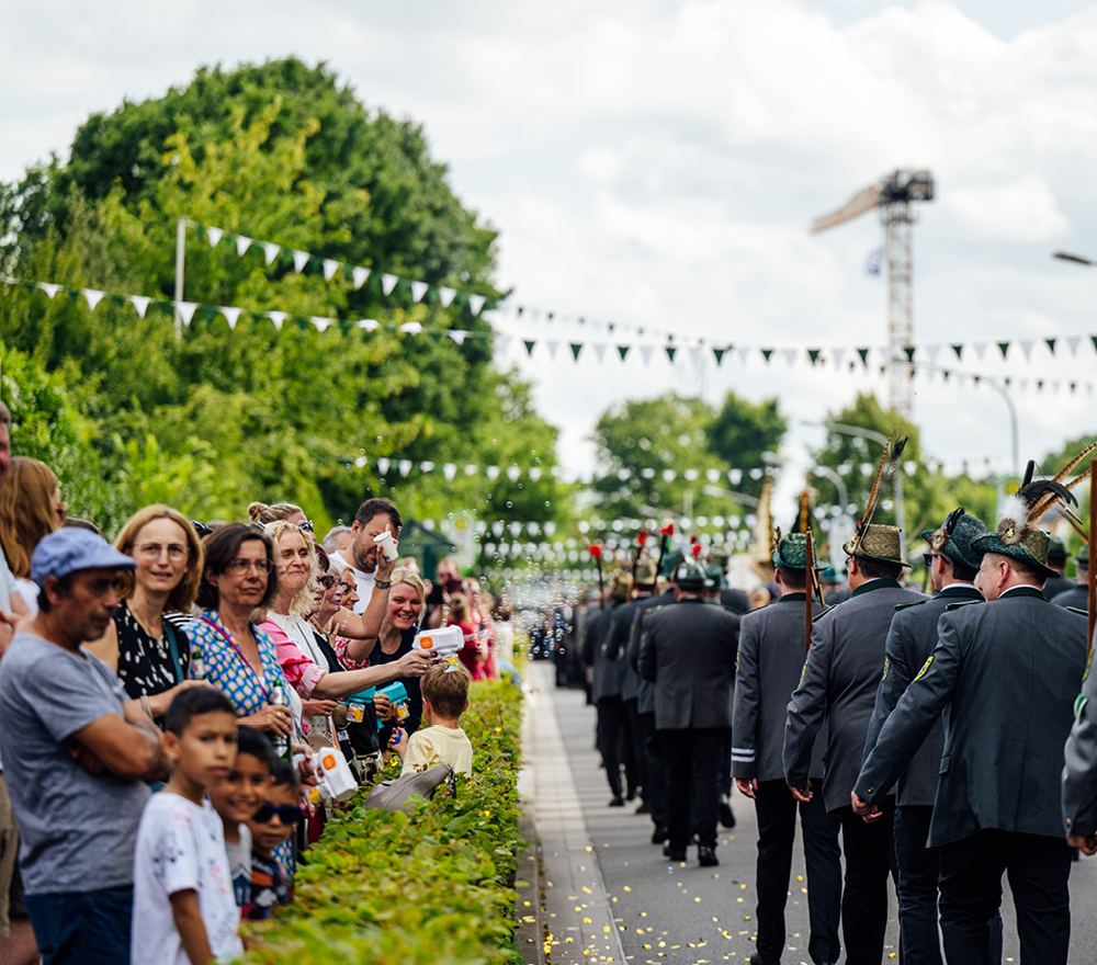 Eine Menschenmenge säumt eine Straße und beobachtet eine Parade von Menschen in dunklen Uniformen und Hüten. Bunte Wimpel hängen über dem Kopf, und einige Kinder stehen in der Nähe der Spitze. Im Hintergrund sind Bäume und ein Kran bei teilweise bewölktem Himmel zu sehen.
