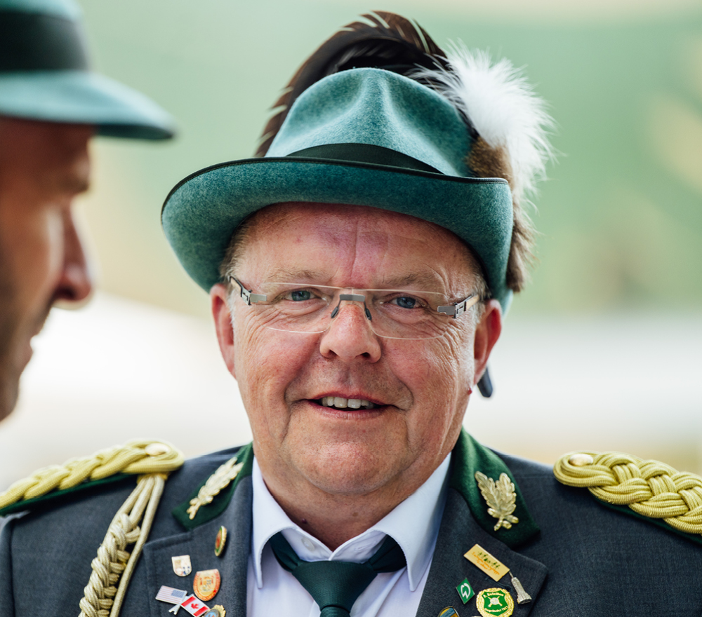 Ein Mann in grüner Trachtenuniform mit goldenen Verzierungen, Anstecknadeln und Abzeichen, der einen grünen Hut mit einer Feder und eine Brille trägt, lächelt in die Kamera. Eine weitere Person mit einem ähnlichen Hut ist teilweise links zu sehen.