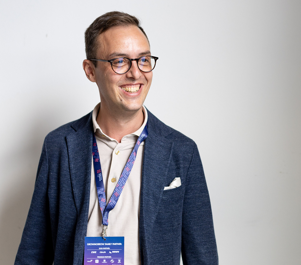 Ein lächelnder Mann, der eine Brille, einen blauen Blazer und ein Schlüsselband mit einem Veranstaltungsabzeichen trägt, steht vor einem schlichten weißen Hintergrund.
