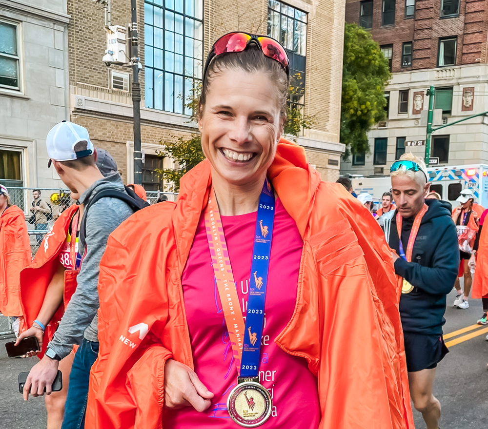 Eine lächelnde Frau, die eine Medaille und eine orangefarbene Laufdecke trägt, steht nach dem Zieleinlauf eines Marathons im Freien zwischen anderen Läufern. Sie trägt eine Laufkleidung und eine Sonnenbrille, im Hintergrund sind Gebäude zu sehen.