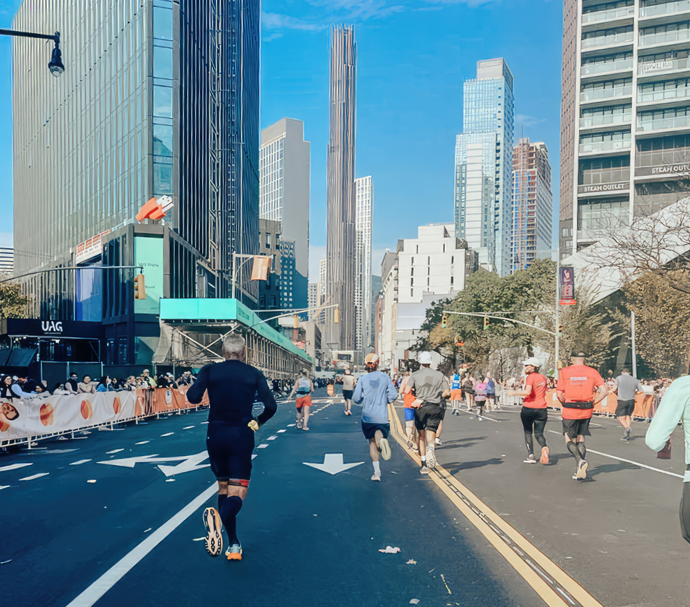 Läufer nehmen an einem Stadtmarathon teil, der an einem sonnigen Tag von hohen modernen Wolkenkratzern umgeben ist. Zuschauer säumen die Straße hinter Absperrungen, und die Fahrspuren sind mit weißen Pfeilen markiert.