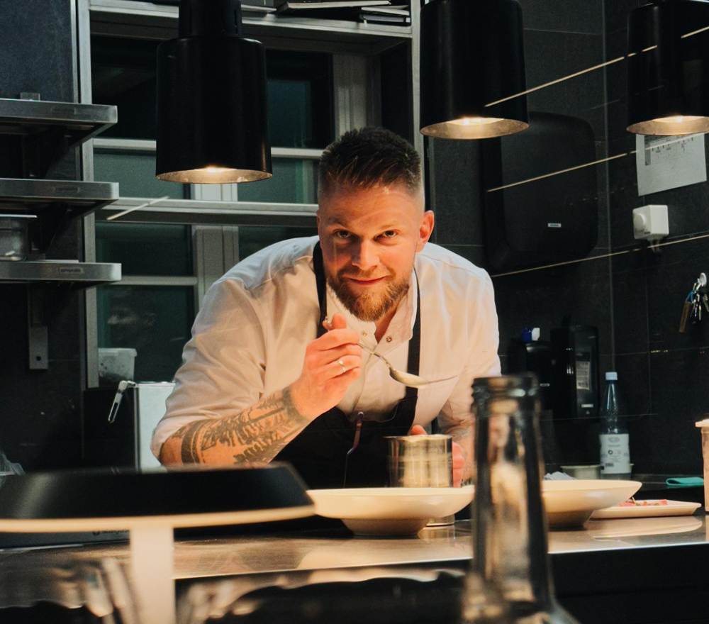 Ein Koch mit Tattoos und Bart, der einen weißen Kittel und eine Schürze trägt, lehnt sich in einer modernen Küche nach vorne, lächelt und hält einen Löffel über ein angerichtetes Gericht, während er unter Hängelampen das Essen zubereitet.