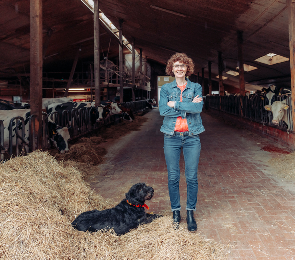 Eine Frau steht lächelnd mit verschränkten Armen in einer Scheune, umgeben von Kühen in Ställen. Sie trägt eine Jeansjacke und Jeans. Ein schwarzer Hund sitzt auf einem Haufen Heu zu ihren Füßen. Die Scheune ist geräumig und hat einen gemauerten Boden und Holzbalken.