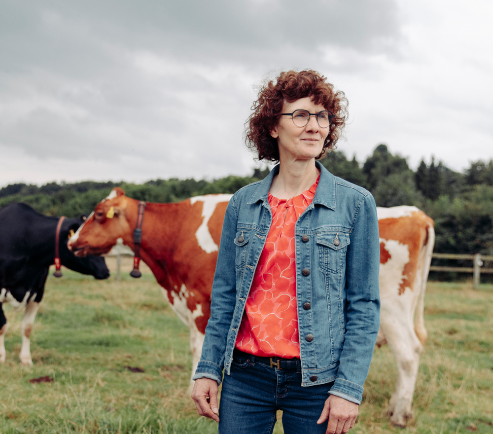 Eine Frau mit lockigem Haar, die eine Brille, eine Jeansjacke und ein leuchtend orangefarbenes Oberteil trägt, steht auf einem grasbewachsenen Feld mit zwei Kühen im Hintergrund. Hinter ihr sind Bäume und ein bewölkter Himmel zu sehen.