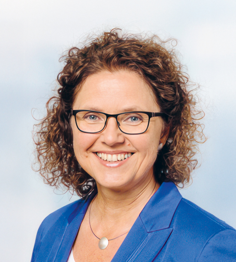 Eine Frau mit lockigem braunem Haar und Brille, die einen blauen Blazer und ein weißes Oberteil trägt, lächelt in die Kamera vor einem hellen Hintergrund.