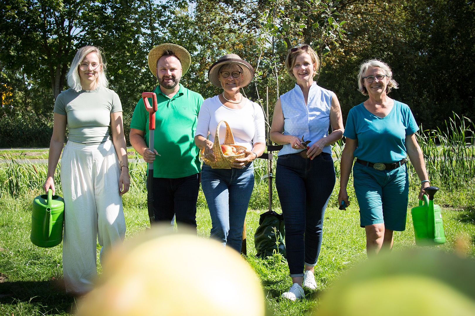 Fünf Erwachsene stehen im Freien in einem sonnigen Garten, lächeln und halten Gartengeräte und Körbe in der Hand. Sie sind von grünem Gras und Bäumen umgeben.