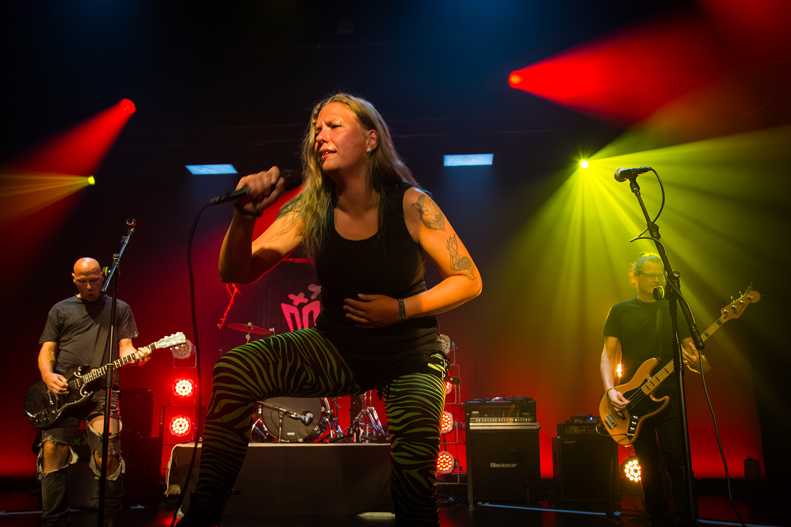 Auf der Bühne singt eine Frau leidenschaftlich in ein Mikrofon. Sie trägt eine schwarz-grüne Hose mit Zebradruck, die an die Ausrüstung von Schlittenrettung im Wattenmeer erinnert. Neben ihr spielen zwei Männer Gitarre und Bass, während im Hintergrund bunte Bühnenlichter leuchten.