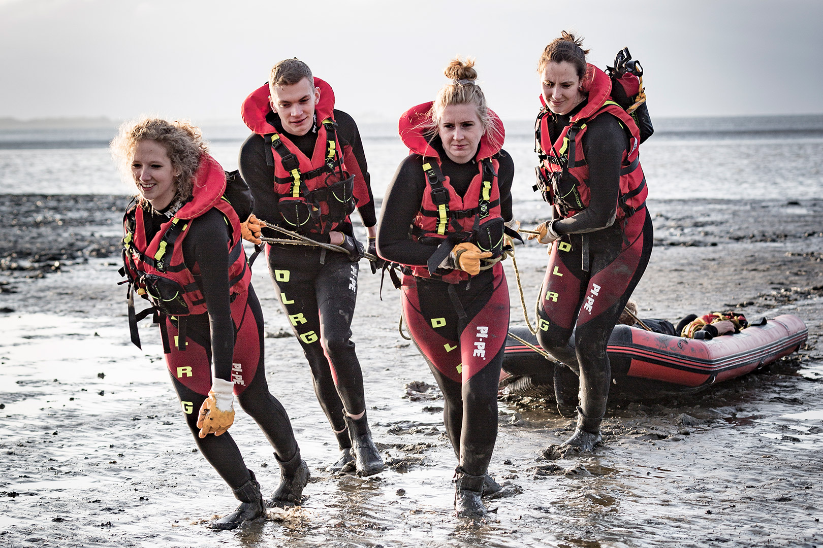 Vier Personen in Neoprenanzügen und roten Schwimmwesten ziehen ein aufblasbares Rettungsboot über ein schlammiges Ufer. Sie wirken konzentriert und arbeiten in der Nähe des Ufers als Team zusammen.