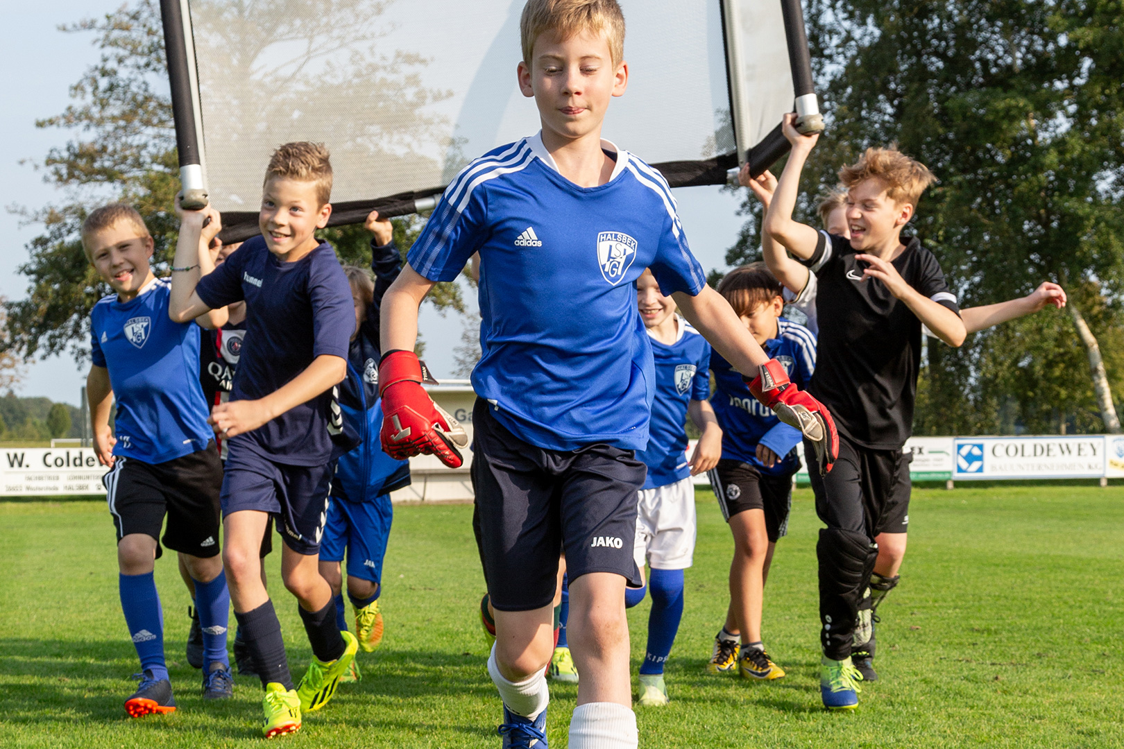 Eine Gruppe lächelnder Kinder in blauen Fußballtrikots trägt einen Torpfosten hinter einem Jungen mit Torwarthandschuhen her und tut spielerisch so, als sei es eine Schlittenrettung im Wattenmeer auf einer Wiese unter sonnigem Himmel.