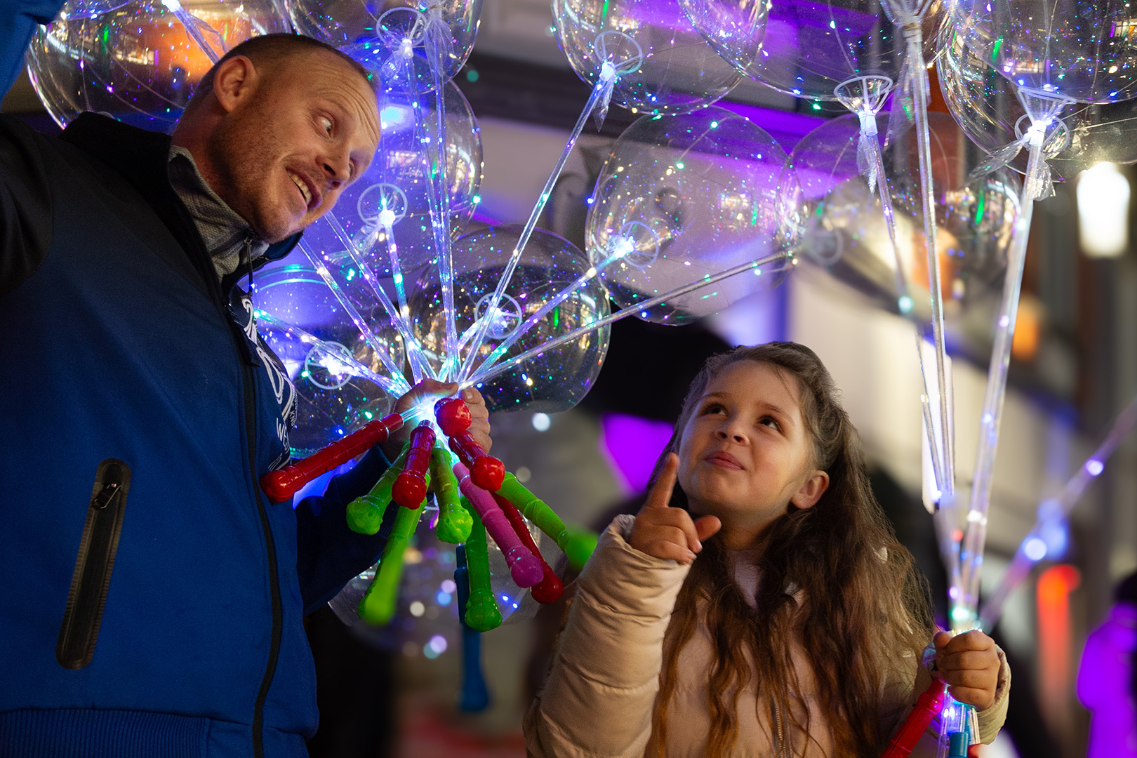 Ein Mann und ein junges Mädchen lächeln und blicken nach oben, während sie leuchtende, bunte LED-Ballons in der Hand halten. Ihre Freude erinnert an die Aufregung während einer Schlittenrettung im Wattenmeer. Die Szene ist festlich, mit hellen Lichtern und einer fröhlichen Atmosphäre.