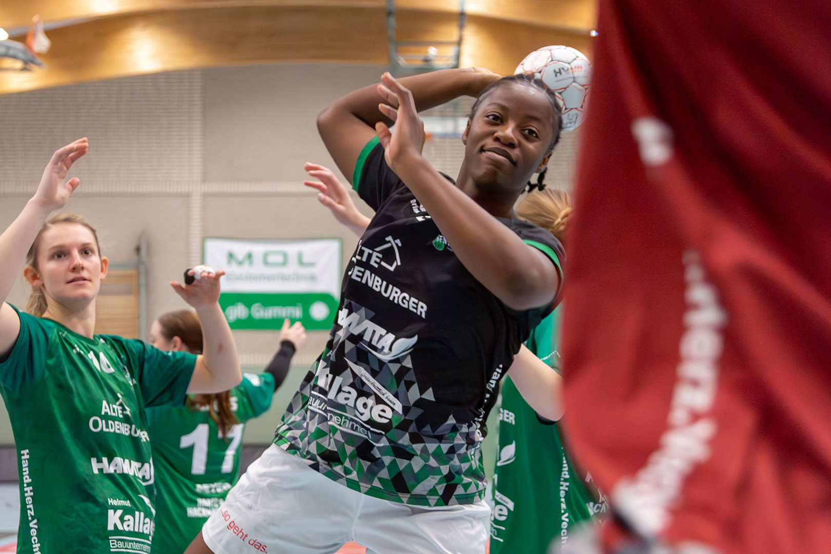 Eine Handballspielerin in einem schwarzen Trikot bereitet sich auf den Wurf vor, während ihre Gegnerinnen in grünen Trikots versuchen, sie zu blocken.