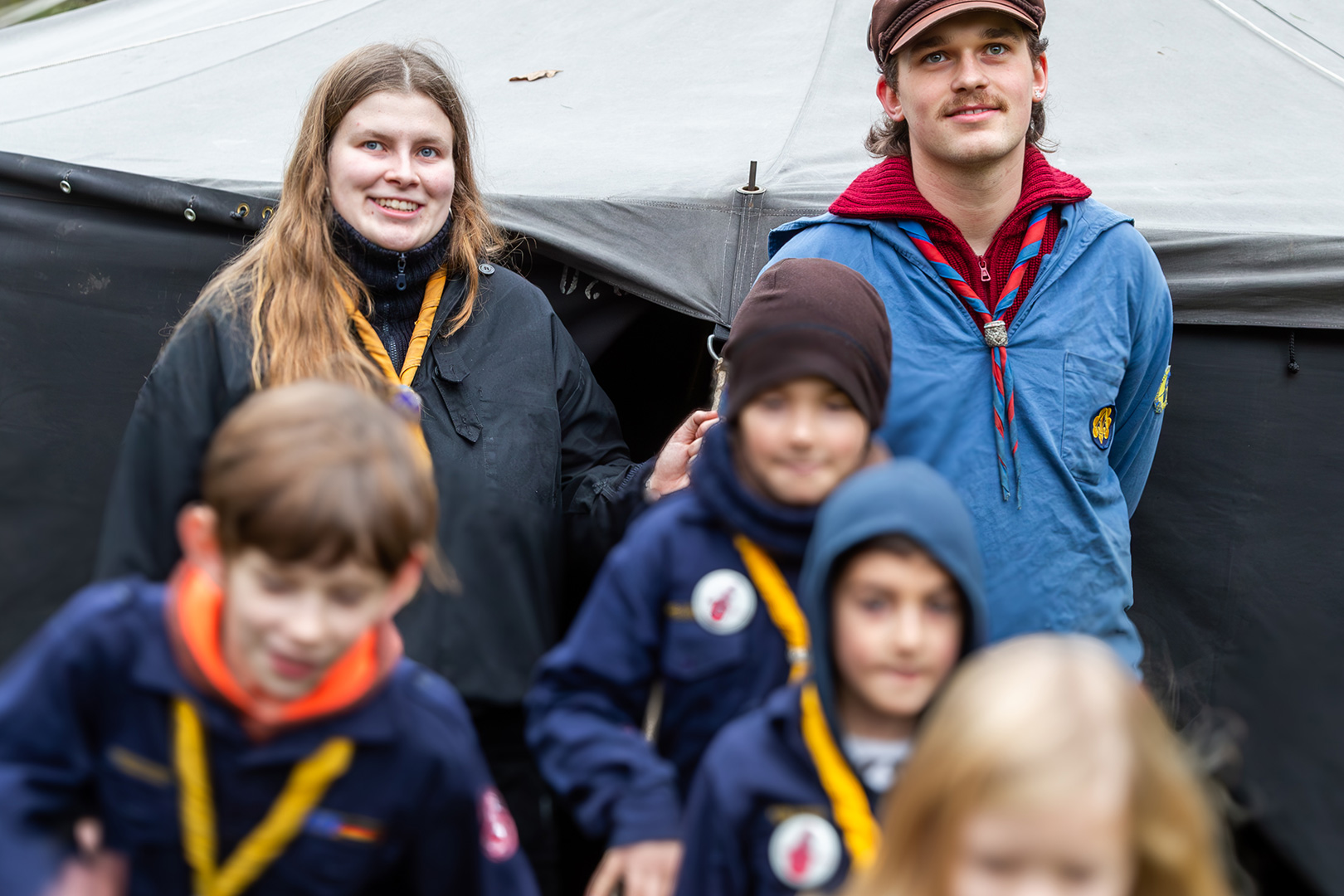 Eine Gruppe von Kindern und zwei Erwachsene in Pfadfinderuniformen mit Halstüchern stehen vor einem großen dunklen Zelt im Freien und bereiten sich vielleicht auf die Schlittenrettung im Wattenmeer vor. Die Erwachsenen lächeln, während die Kinder im Vordergrund stehen oder sich bewegen.