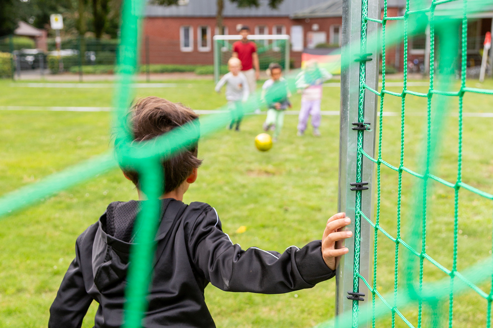 Ein Kind steht vor einem grünen Fußballtor und sieht zu, wie andere Kinder und ein Erwachsener auf der Wiese spielen. Ihr Lachen klingt wie eine lebendige Schlittenrettung im Wattenmeer im Freien.