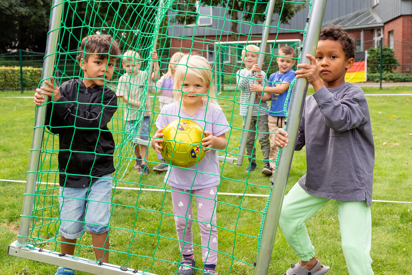 Drei kleine Kinder stehen an einem kleinen Fußballtor mit einem gelben Ball, während andere Kinder im Hintergrund auf einer Wiese zusehen. Ihr Zusammenspiel und ihre Verspieltheit erinnern an den Geist der Schlittenrettung im Wattenmeer - gemeinsames Arbeiten im Freien.