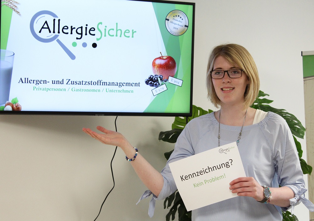 Eine Frau steht neben einem Bildschirm, auf dem AllergieSicher und Informationen über den Umgang mit Allergenen und Zusatzstoffen angezeigt werden. Sie hält ein Schild mit der Aufschrift Kennzeichnung? Kein Problem! und gestikuliert in Richtung des Bildschirms.