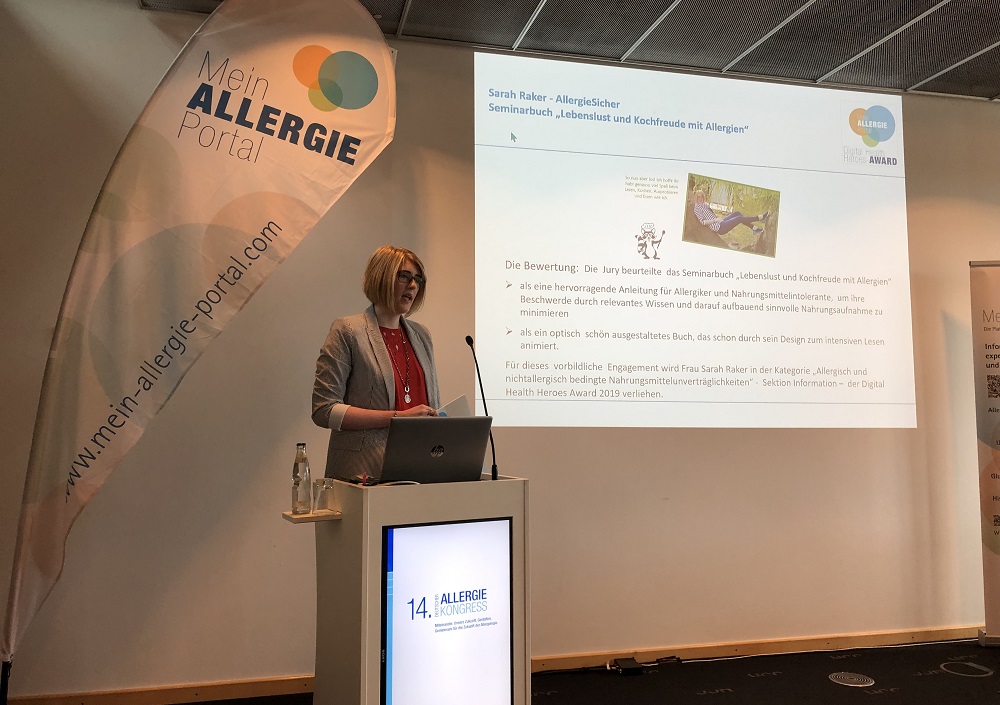 Eine Frau steht an einem Podium und hält einen Vortrag auf dem 14. Allergiekongress. Auf einem Bildschirm hinter ihr ist eine Folie über Allergene zu sehen, und auf einem Banner steht Mein ALLERGIE Portal. Ein Laptop und ein Glas Wasser stehen auf dem Podium.