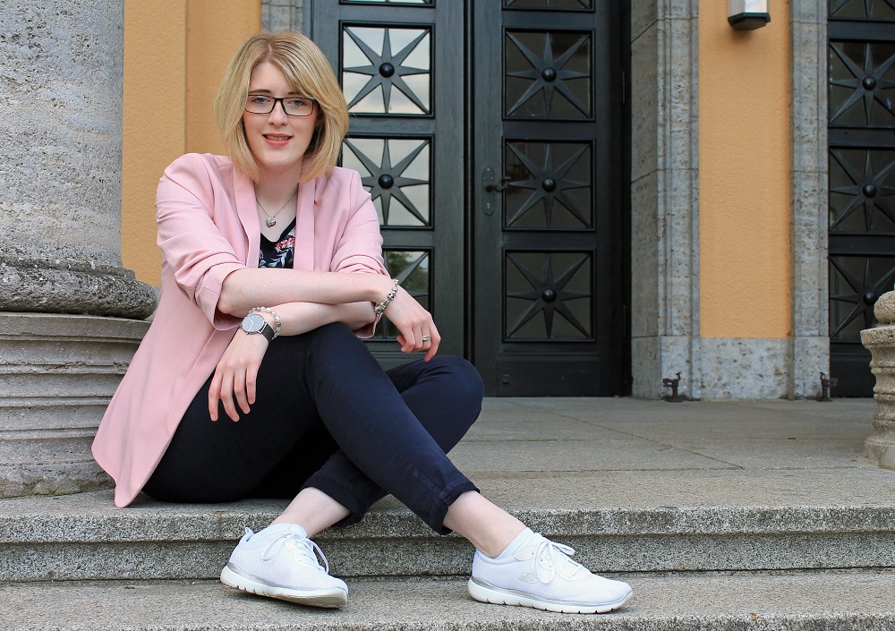 Eine junge Frau mit blondem Haar, Brille und rosa Blazer sitzt lässig auf den Steinstufen vor einer großen schwarzen Doppeltür und lächelt in die Kamera.