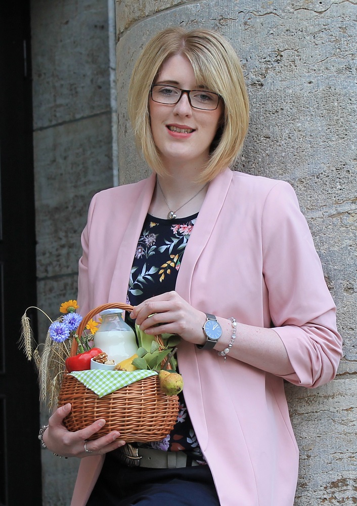 Eine Frau mit blondem Haar, Brille und rosafarbenem Blazer steht an einer Steinmauer, lächelt und hält einen kleinen Korb mit buntem Gemüse, Blumen und einer Flasche in der Hand.