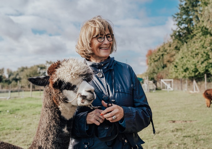 Eine lächelnde Frau mit Brille und blauer Jacke steht neben einem braun-weißen Alpaka in einem grasbewachsenen Außenbereich mit Zäunen und Bäumen im Hintergrund.
