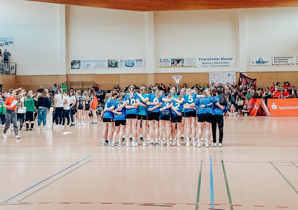 Ein Mädchensportteam in blauen Trikots steht in einer Turnhalle zusammen, während sich im Hintergrund andere Teams und Zuschauer versammeln. Die Szene scheint während einer Hallensportveranstaltung oder eines Turniers zu spielen.