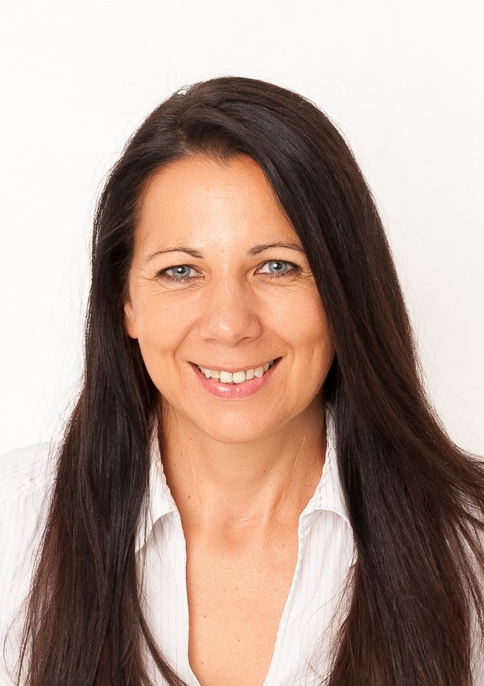 Eine Frau mit langen dunklen Haaren und blauen Augen lächelt in die Kamera. Sie trägt ein weißes Hemd mit Kragen und posiert vor einem schlichten weißen Hintergrund.