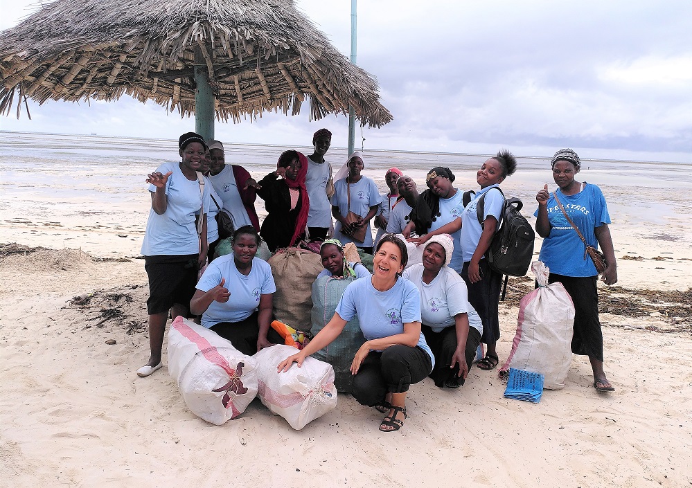 Eine Gruppe von Menschen in passenden blauen Hemden posiert und lächelt an einem Strand unter einem strohgedeckten Sonnenschirm mit großen Säcken gesammelten Mülls, um Teamwork bei einer Strandsäuberungsaktion zu zeigen.