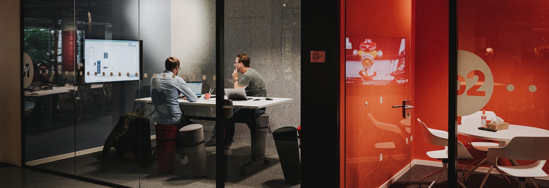 Zwei Personen sitzen an einem Tisch in einem gläsernen Besprechungsraum mit Laptops und schauen auf einen Bildschirm. Neben ihnen ist ein weiterer leerer Besprechungsraum mit einer roten Wand und einem runden Tisch zu sehen.