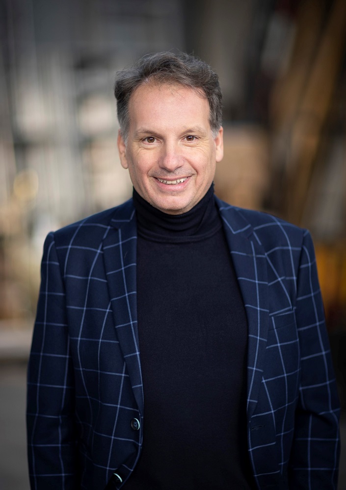 Ein Mann mit kurzen grauen Haaren, der einen marineblau karierten Blazer und einen schwarzen Rollkragenpullover trägt, lächelt in die Kamera vor einem unscharfen Hintergrund.