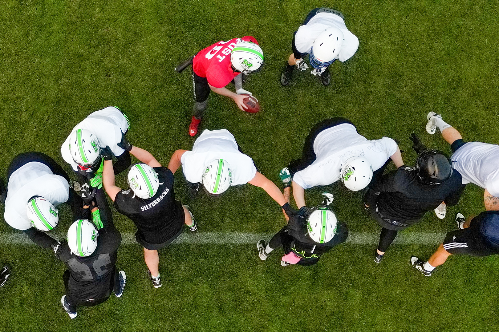 Luftaufnahme einer Fußballmannschaft in weißen Trikots, die eine Reihe bildet, während ein Spieler in einem roten Trikot einen Fußball hält und sich auf ein Spiel auf einem grünen Feld vorbereitet, was an die Teamarbeit bei einer Schlittenrettung im Wattenmeer erinnert.