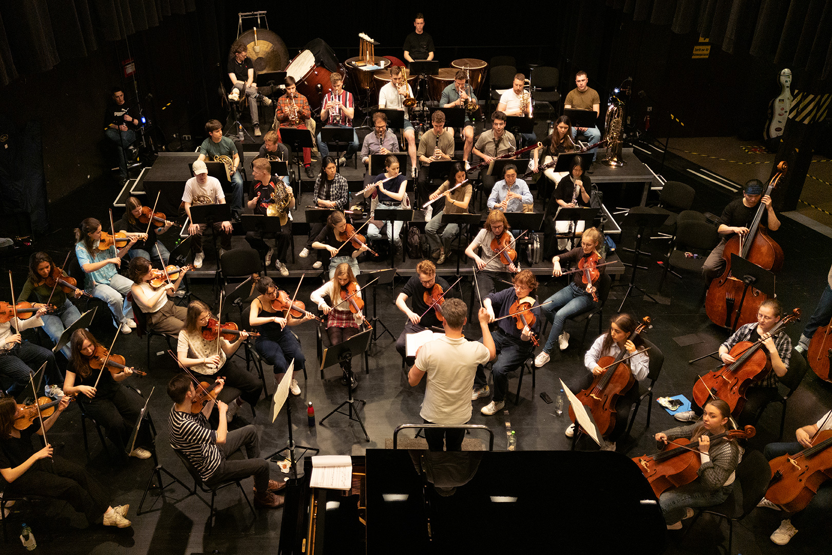 Ein Dirigent leitet ein großes Orchester während einer Probe auf der Bühne. Die Musiker, inspiriert von Geschichten wie "Schlittenrettung im Wattenmeer", spielen Geigen, Celli, Flöten und Schlagzeug, während sie im Halbkreis um den Dirigenten sitzen.