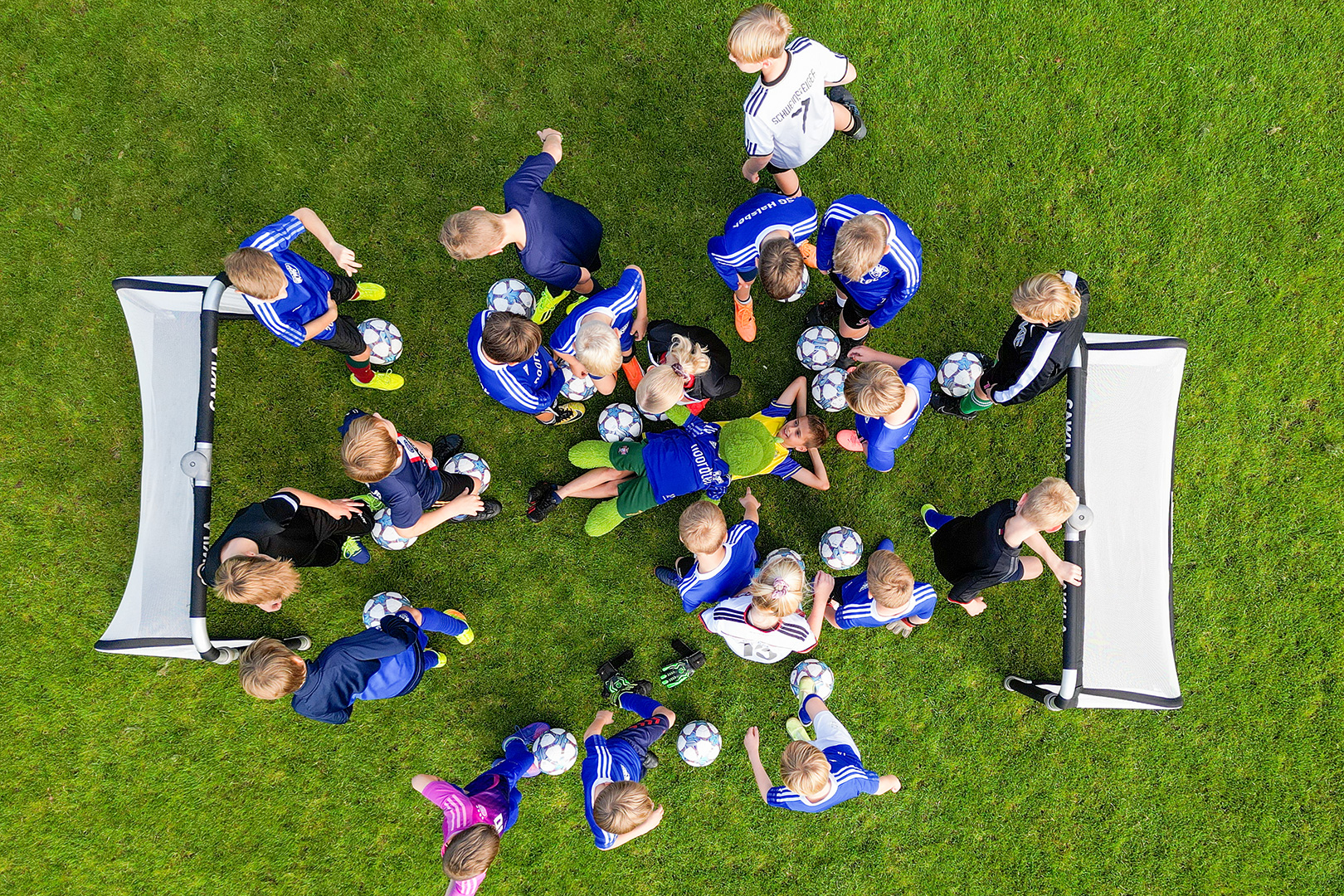 Eine Gruppe von Kindern in Fußballtrikots steht auf dem grünen Rasen zwischen zwei kleinen Toren, jedes hält einen Fußball in der Hand, von oben betrachtet - einsatzbereit wie eine Mannschaft der Schlittenrettung im Wattenmeer, die sich auf die Rettung vorbereitet.