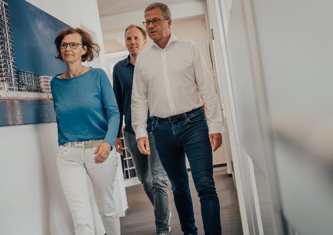 Drei Erwachsene, zwei Männer und eine Frau, gehen gemeinsam einen Flur entlang. Die Frau trägt ein blaues Oberteil und eine weiße Hose, während die Männer helle Hemden und Jeans tragen. An der Wand hängt ein Foto von einem Gebäude am Wasser.
