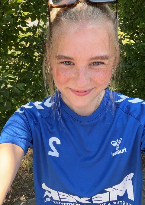 Eine junge Frau mit blondem Haar, die eine Sonnenbrille auf dem Kopf und ein blaues Sporttrikot mit der Nummer 2 trägt, lächelt in die Kamera, während sie draußen vor grünen, belaubten Sträuchern steht.